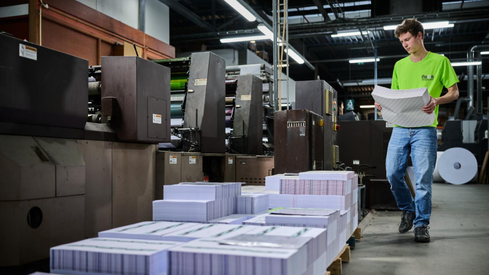 Employee stacking printed materials beside industrial printing presses in a production facility